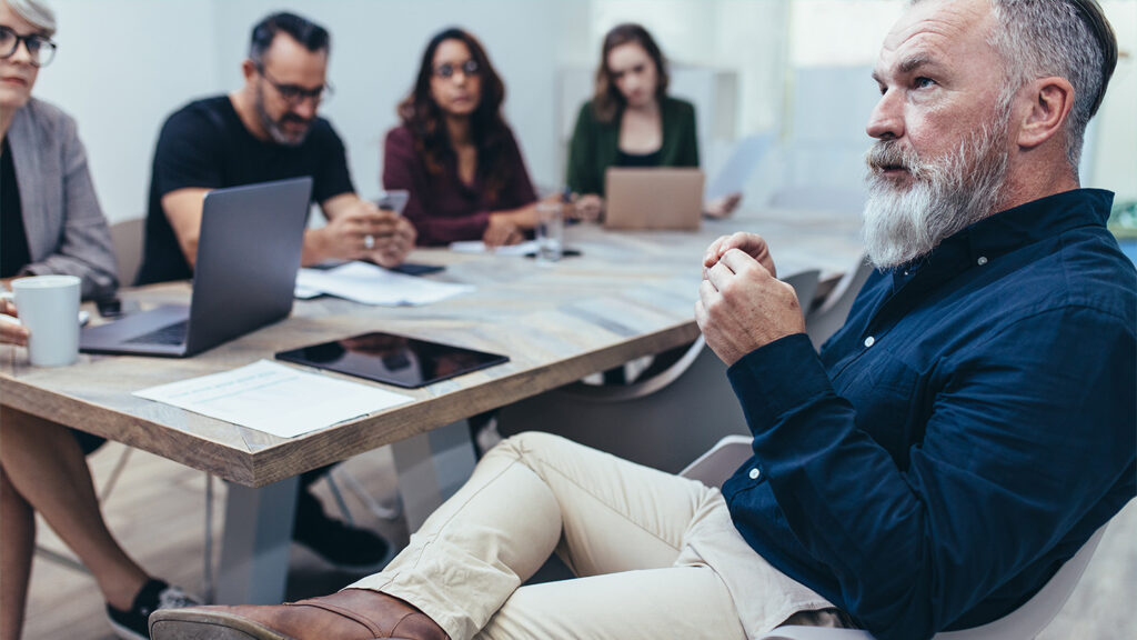 Eine Gruppe Menschen sitzt an einem hellen Konferenztisch aus Holz. Im Vordergrund ist ein Mann in blauem Hemd und heller Hose zu sehen. Er trägt einen längeren weiß-grauen Bart. Die anderen Personen im Hintergrund sind unscharf zu erkennen. Der Mann hat das eine Bein über das andere geschlagen und schaut konzentriert auf etwas Außerhalb des Bildes.