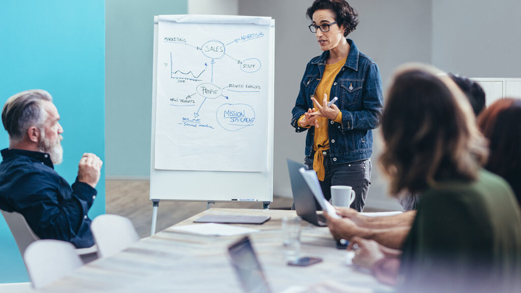 Eine Frau mit dunklen Haaren, einem gelben Shirt und einer blauen Jeansjacke steht neben einem Flipchart in einem Konferenzraum. Eine Gruppe Menschen sitzt an einem hellen Konferenztisch und hat den Blick auf die Frau gerichtet. Die Gruppe hört konzentriert zu, während die Frau etwas erklärt.