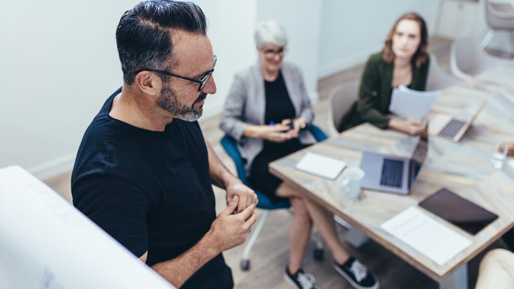 Ein Mann mit dunklen Haaren und einem dunklen T-Shirt steht vor einem hellen Konferenztisch aus Holz. Zwei Frauen sitzen an dem Konferenztisch und hören ihm konzentriert zu. Die beiden sind unscharf im Hintergrund zu erkennen. Auf dem Tisch liegen Dokumente, Laptops und ein Glas und eine Kaffeetasse.