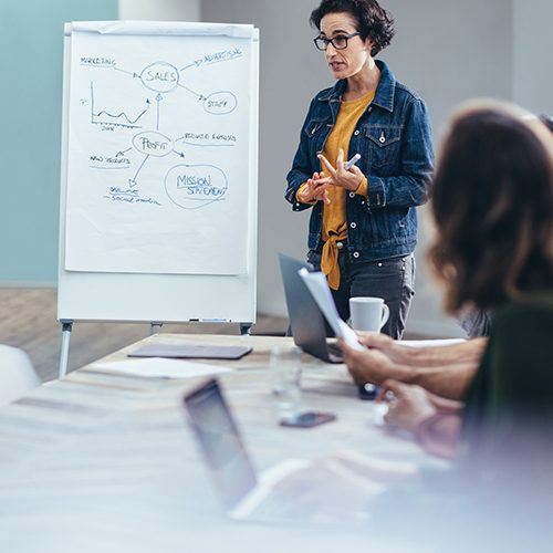 Eine Frau mit dunklen Haaren, einem gelben Shirt und einer blauen Jeansjacke steht neben einem Flipchart in einem Konferenzraum. Eine Gruppe Menschen sitzt an einem hellen Konferenztisch und hat den Blick auf die Frau gerichtet. Die Gruppe hört konzentriert zu, während die Frau etwas erklärt.